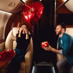 Romantické prekvapenie vo vyhliadkovom lietadle – muž daruje žene so srdiečkom darček a kyticou ruží.